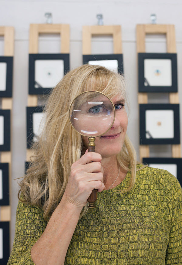 Artist Karen Libecap Holding a Magnifying Glass while standing in front of a wall of her miniature artwork.